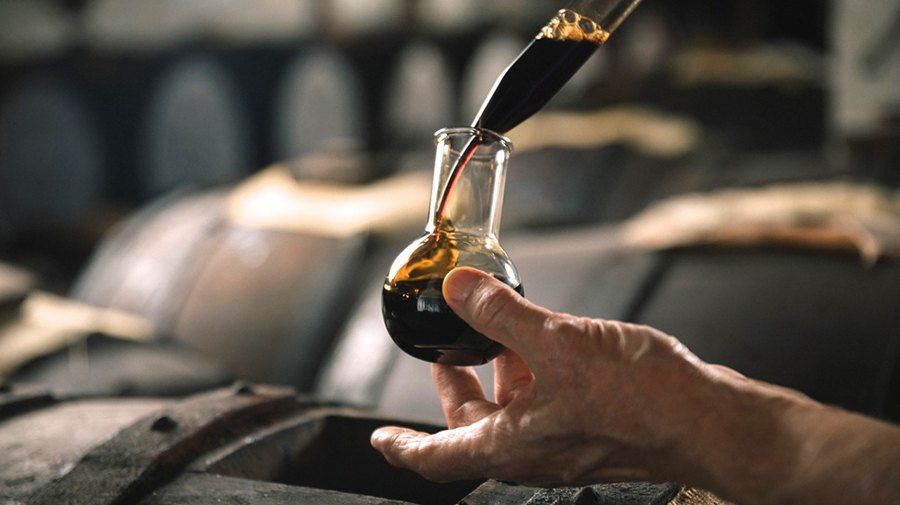 Dunkler Balsamico, aus einer Glaspipette in ein kleines Fläschchen gegossen, das eine Hand über den Fässern einer Acetaia hält