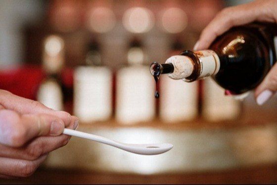 Ein Tropfen gereifter Balsamico, der aus einer verkorkten Flasche auf einen weißen Probierlöffel fällt