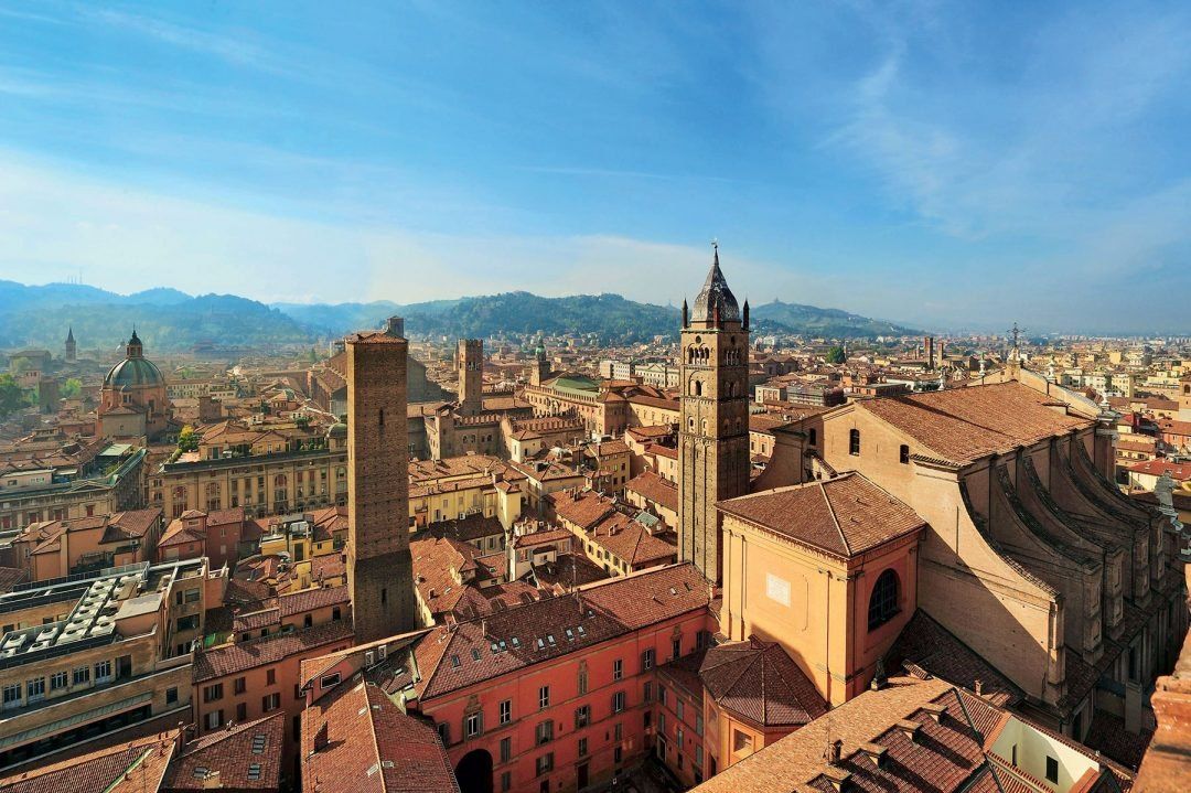 Bolognas Altstadt von oben: mittelalterliche Türme, ein spitzer Glockenturm und Terrakottadächer, dahinter grüne Hügel