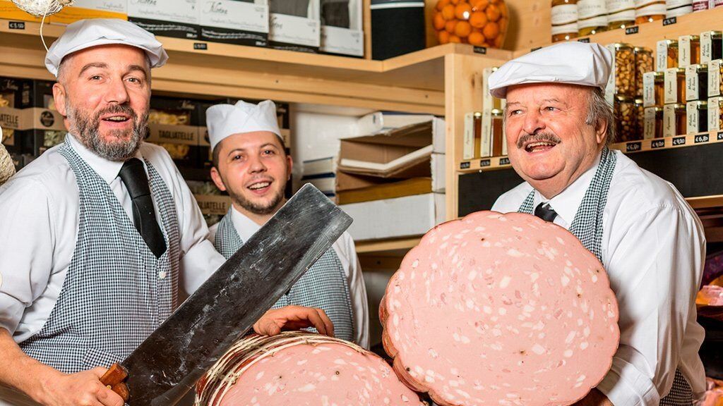 Drei Männer mit weißen Mützen und karierten Schürzen in einer Salumeria, einer mit einem Hackbeil in der Hand, dazu eine ganze Mortadella und eine riesige Scheibe
