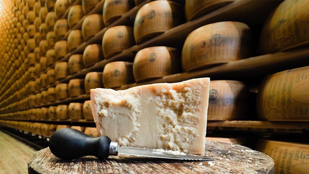 Ein Stück Parmigiano Reggiano und ein Käsemesser auf einem Holzblock, dahinter reifende Laibe, übereinander gestapelt