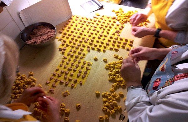 Mehrere Paar Hände beim Formen von Tortellini auf einem Holzbrett, daneben Reihen kleiner Quadrate aus Eierteig mit je einem Tupfer Füllung und eine Schüssel mit Füllung