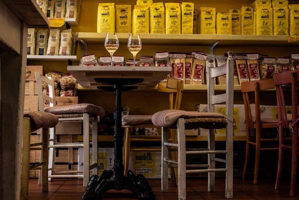 Zwei Gläser Weißwein auf einem kleinen Trattoria-Tisch, dahinter Regale mit getrockneter Pasta