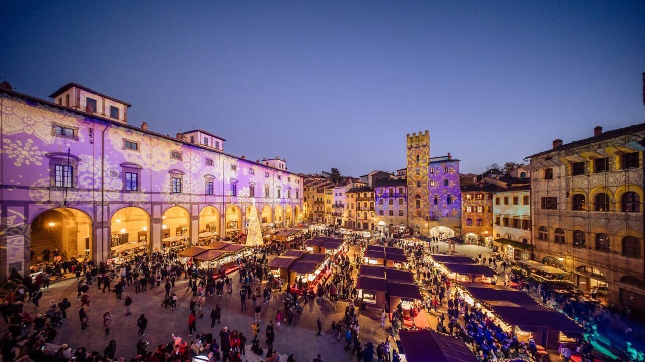 Der Weihnachtsmarkt auf der Piazza Grande in Arezzo in der Abenddämmerung, mit auf die Logge del Vasari projizierten Lichtmustern