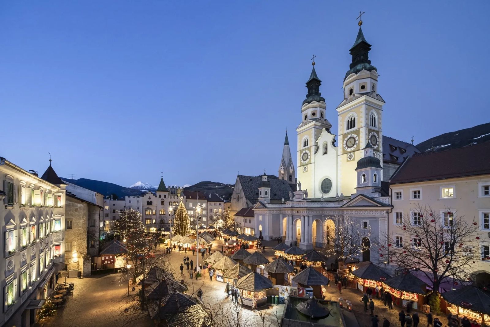 Stände des Weihnachtsmarkts auf dem Domplatz von Brixen (Bressanone) in Südtirol in der Abenddämmerung, dahinter verschneite Gipfel
