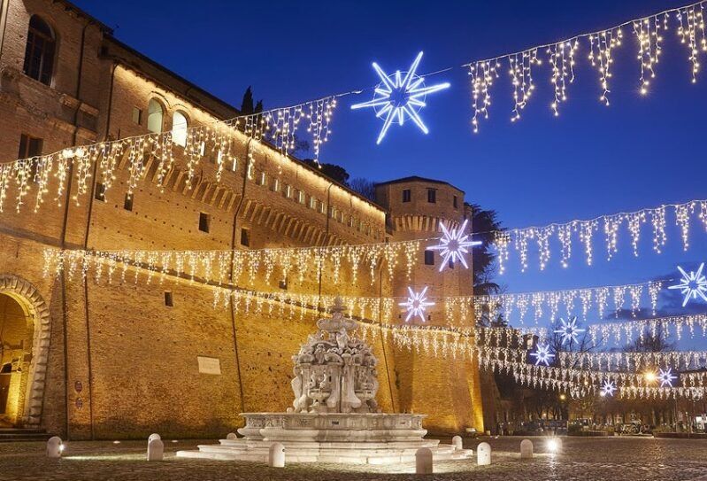 Weihnachtliche Lichterketten und Sterne über einer Piazza in der Abenddämmerung, vor einer Festung aus Backstein und einem Brunnen aus gemeißeltem Marmor