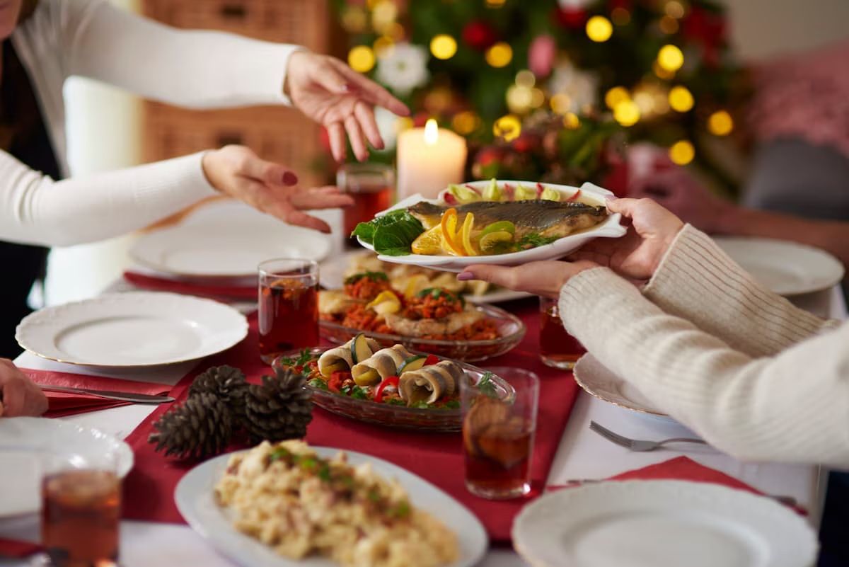 Eine Fischplatte wird über einen festlich gedeckten Weihnachtstisch gereicht, dahinter Kerzen und ein beleuchteter Baum