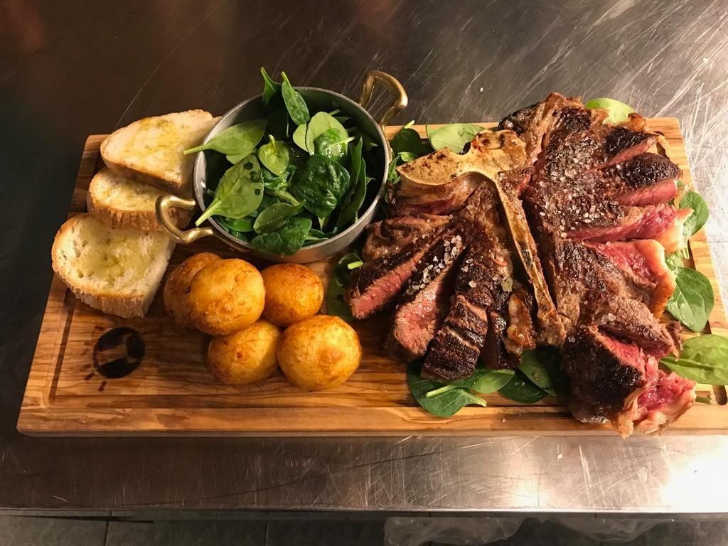 Bistecca alla fiorentina sliced on a wooden board, with golden potatoes, spinach leaves and toasted bread