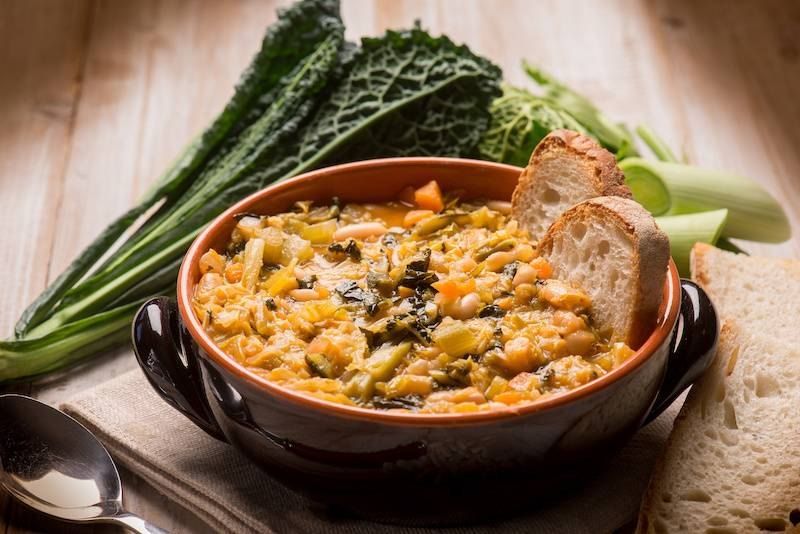 A bowl of ribollita with slices of toasted bread, cavolo nero and fresh vegetables behind