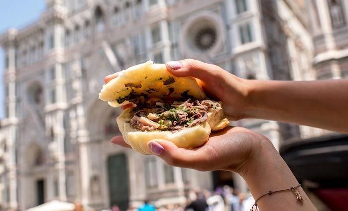 A tripe sandwich held up in front of the marble façade of Florence’s cathedral