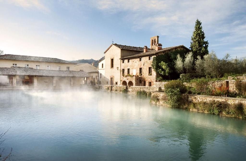 Steam rising from the thermal pool of Bagno Vignoni, lined with old stone buildings and a tall cypress