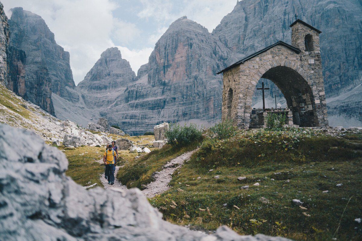 Two hikers on a mountain path near a small stone chapel, beneath the peaks of the Brenta Dolomites