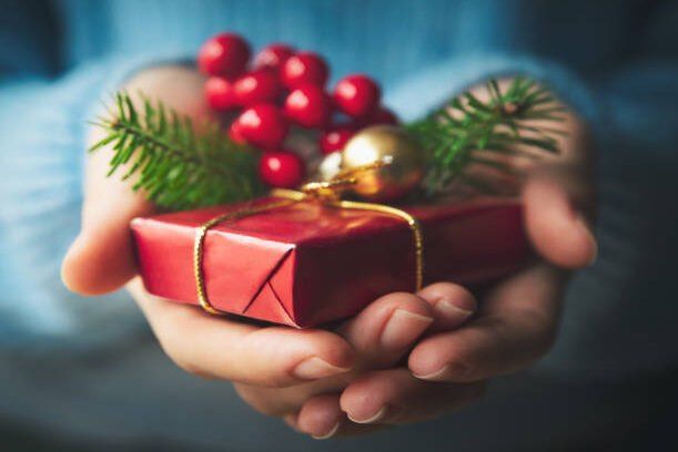 Cupped hands holding a small red present tied with gold ribbon, with red berries, a gold bauble and sprigs of fir
