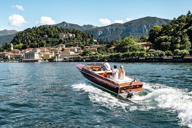 A wooden motorboat carrying three passengers across Lake Como, a lakeside village and wooded mountains behind