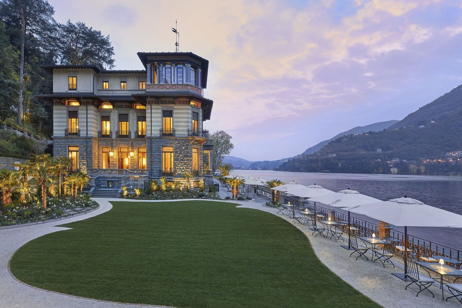 A lakeside villa hotel lit up at dusk on Lake Como, with a lawn and a terrace of parasols along the water
