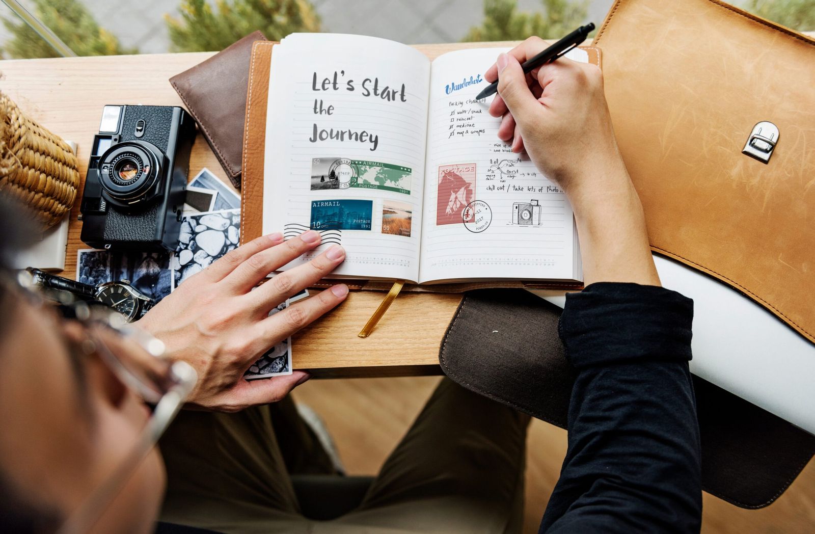 Hands writing in a travel journal, beside a camera, a wristwatch and printed photographs