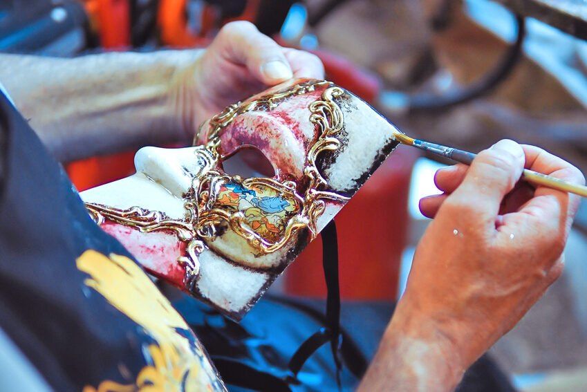 Hands painting gold scrollwork on a Venetian mask with a fine brush