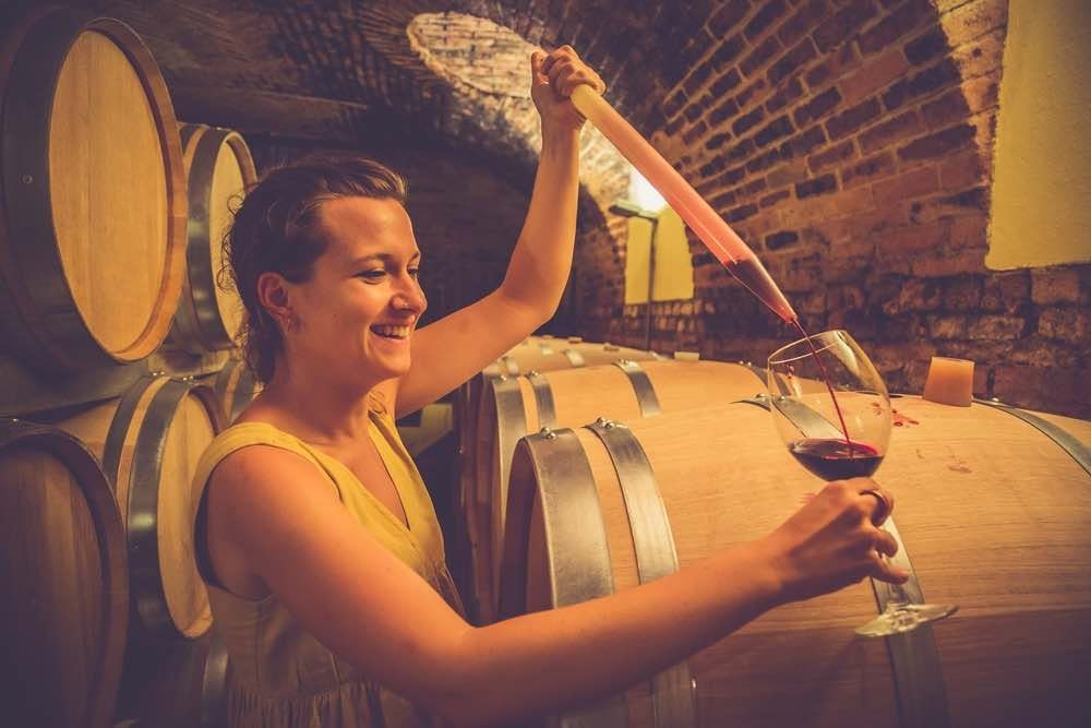 A woman letting red wine run from a wine thief into her glass, among oak barrels in a brick-vaulted cellar
