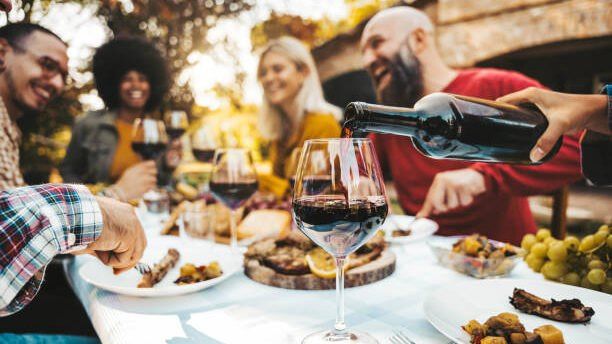 Friends laughing around an outdoor table as red wine is poured into a glass