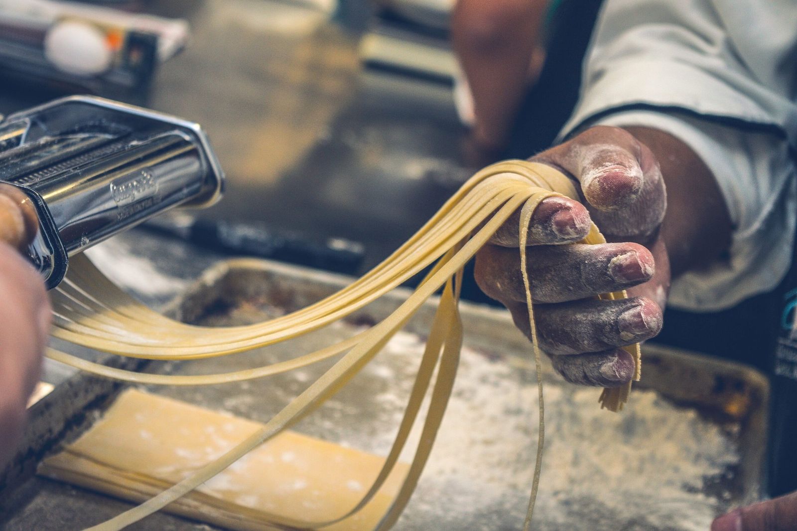A floured hand drawing ribbons of fresh pasta from a pasta machine