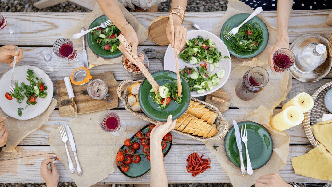 Hands reaching across an outdoor wooden table laid with salads, bread, tomatoes and red wine, seen from above