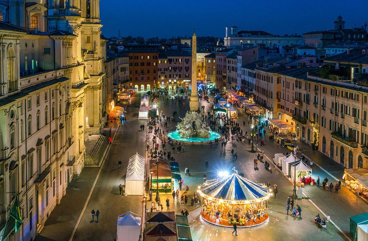 Die Piazza Navona in Rom in der Abenddämmerung während des Weihnachtsmarkts: weiße Marktstände, ein beleuchtetes Karussell und Berninis Vierströmebrunnen