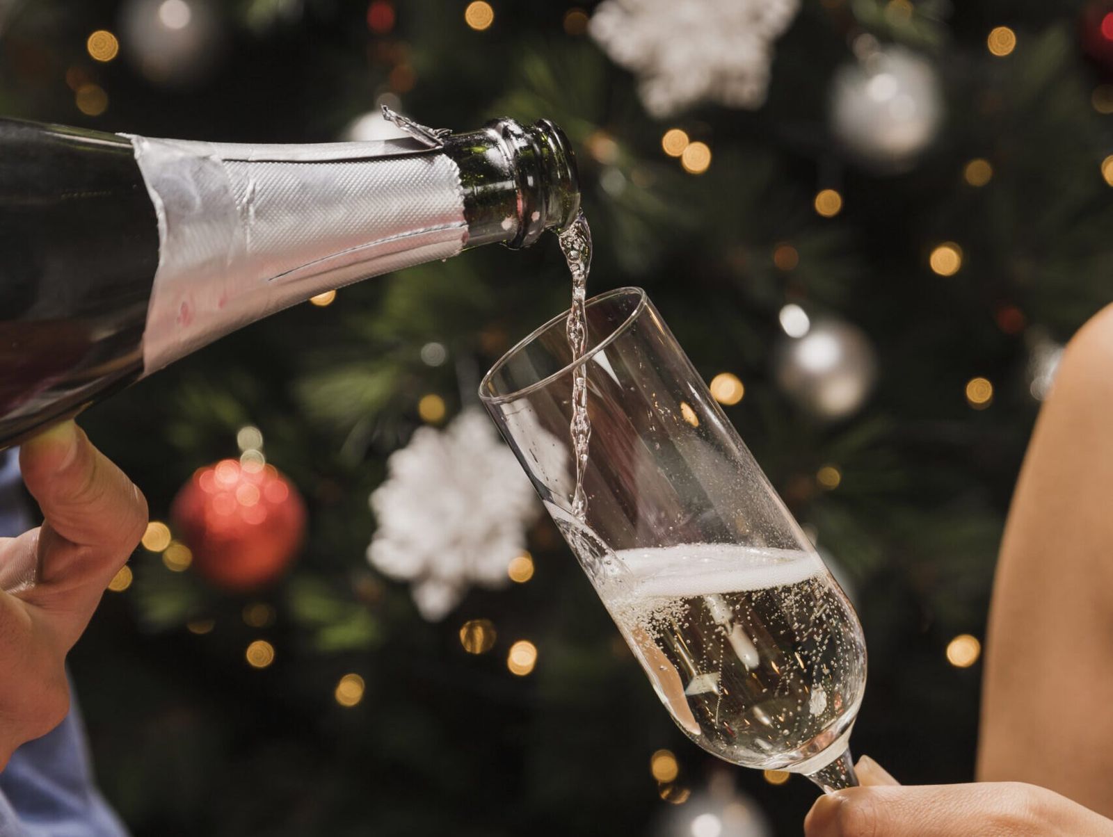 Schaumwein, der vor einem mit Kugeln und Lichtern geschmückten Weihnachtsbaum in ein Sektglas fließt