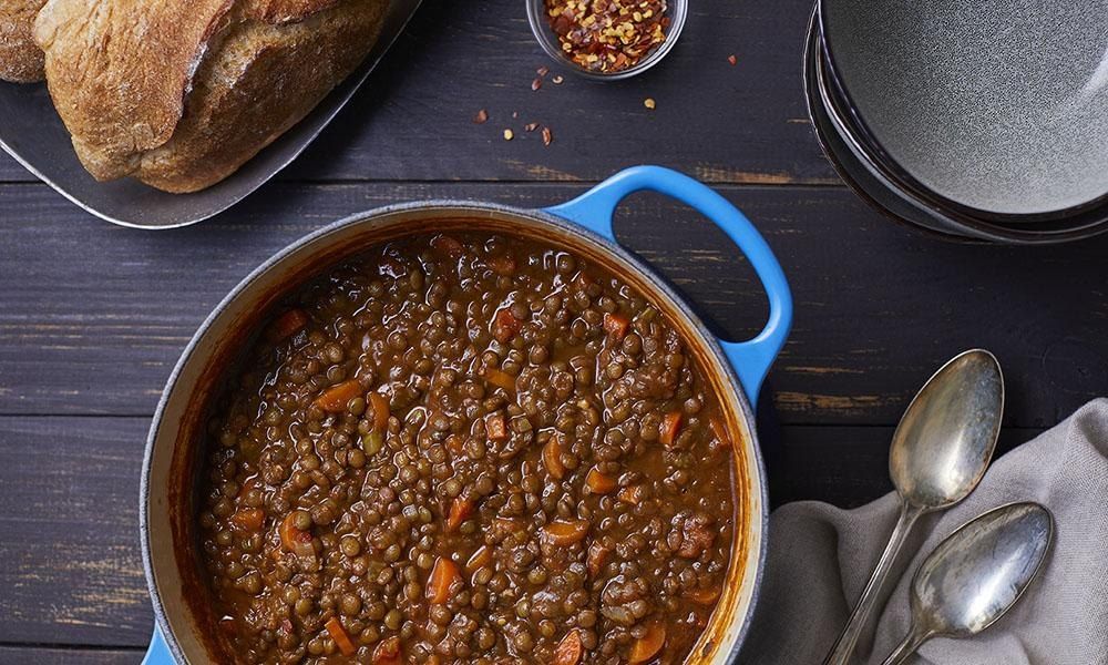 A blue cast-iron pot of lentil soup with carrots, beside a loaf of crusty bread, chilli flakes and two silver spoons