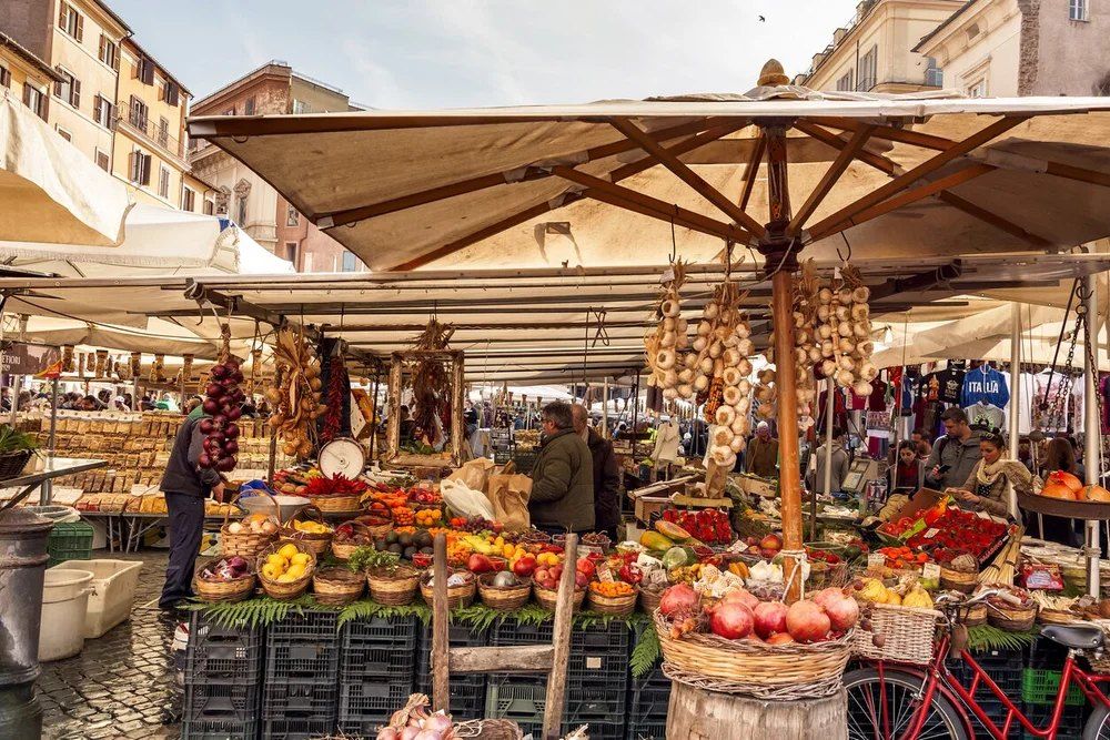 Fruit and vegetable stalls under canvas umbrellas in a cobbled piazza, strings of garlic and onions hanging overhead