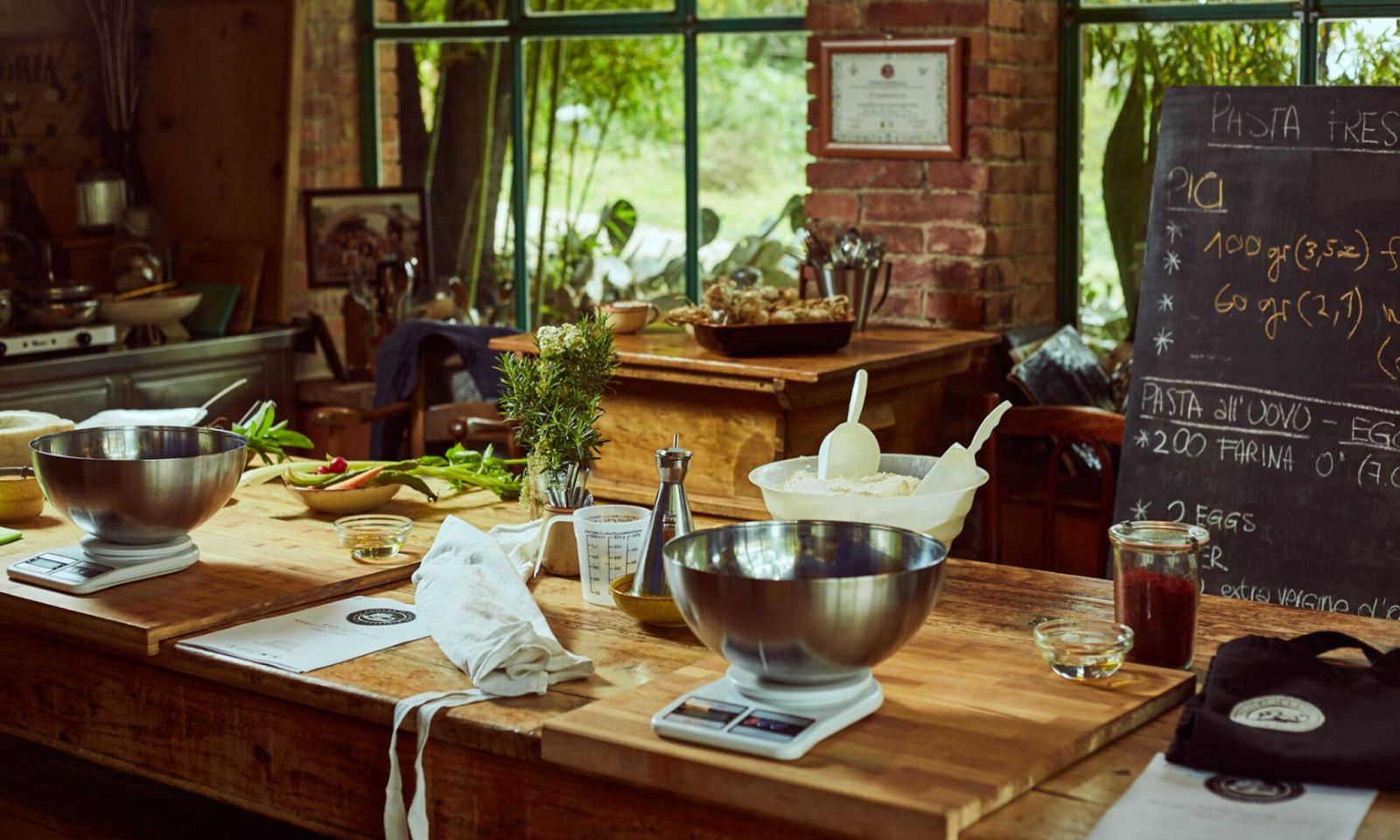 A wooden table laid for a fresh pasta class – scales, steel bowls, flour and rosemary – with the recipe for pici chalked on a blackboard