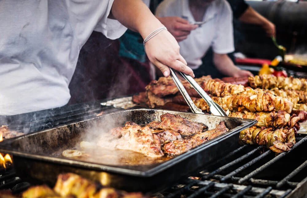 A cook turning meat and skewers with tongs on a smoking grill at a street-food stall