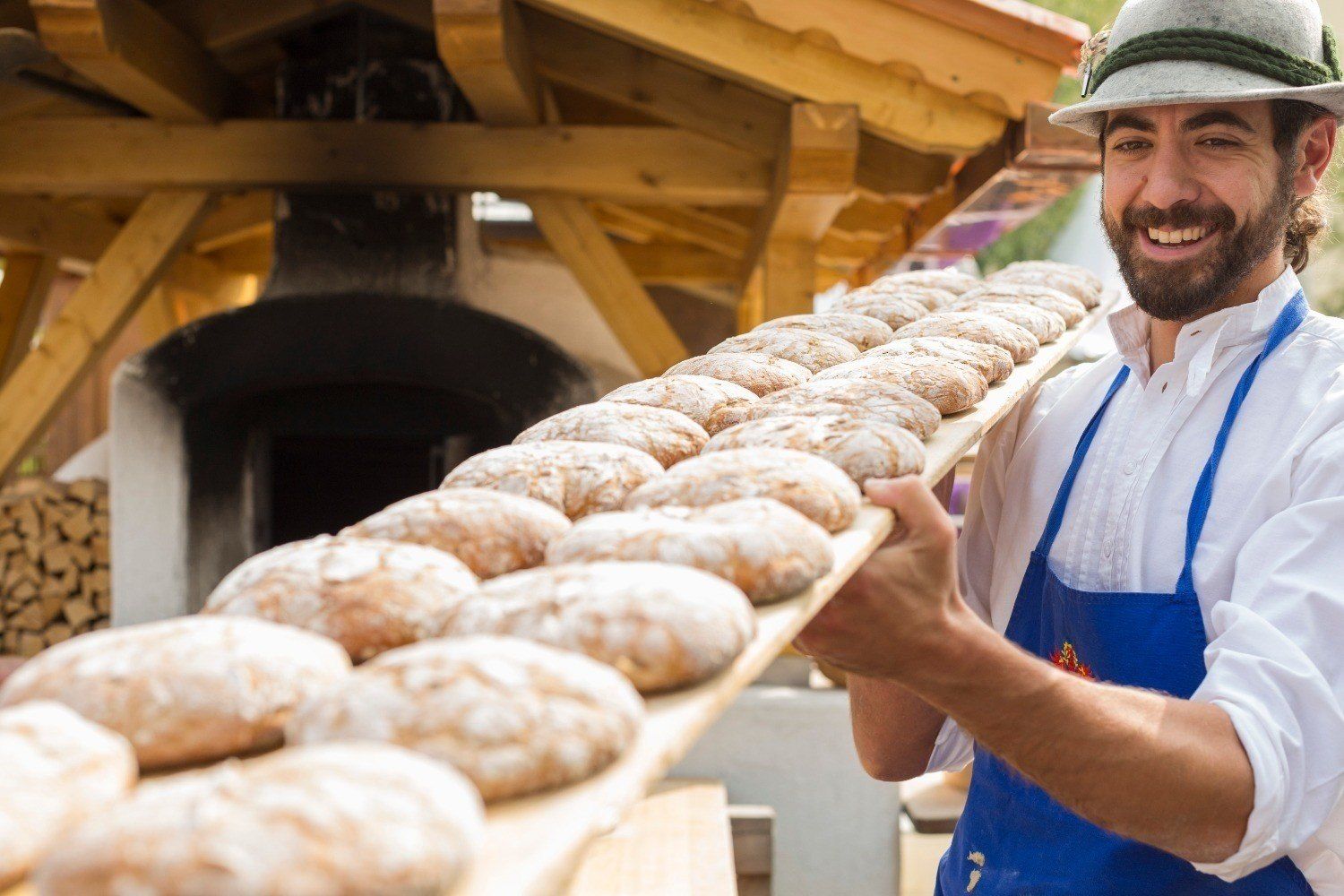 A baker in a felt hat carries a long board of round loaves from a wood-fired oven at the bread and strudel market in Bressanone