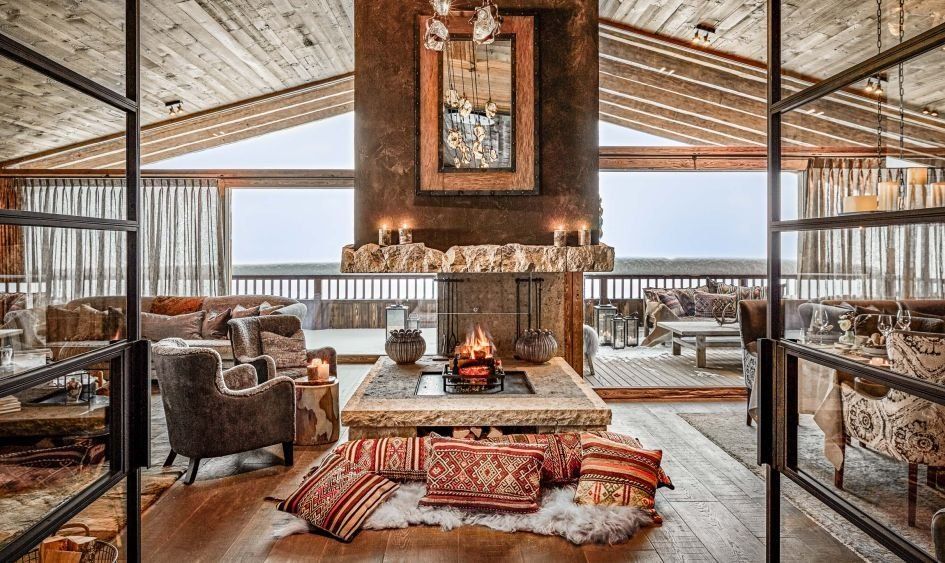 A chalet sitting room with a central open fire, a timber ceiling, armchairs and patterned cushions on a sheepskin rug