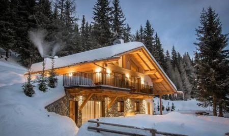 A stone and timber chalet in deep snow among fir trees at dusk, its windows lit and smoke rising from the chimney