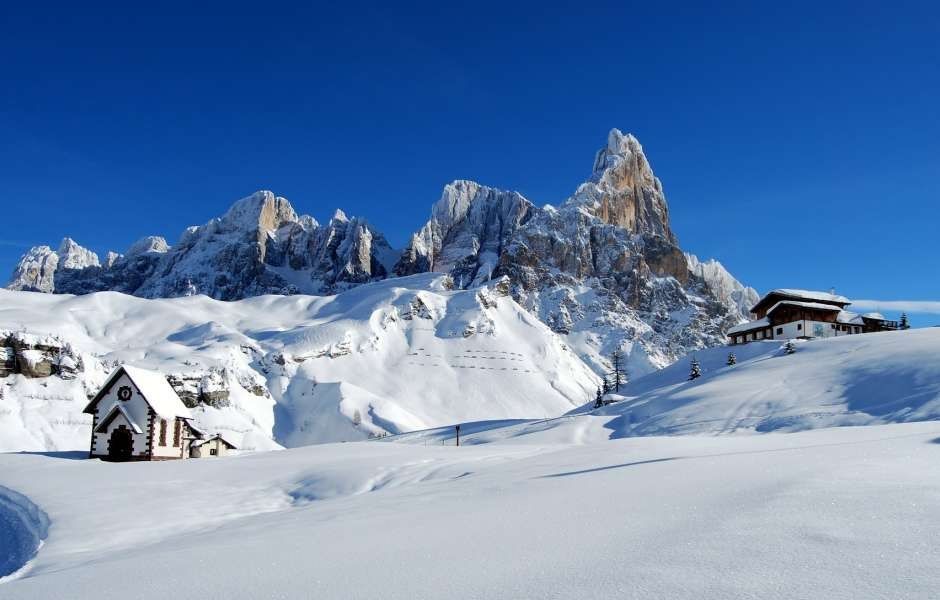 A small chapel and a mountain hut in deep snow beneath jagged Dolomite peaks and a clear blue sky