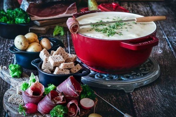 A pot of cheese fondue with bread cubes, small potatoes, broccoli, radishes and rolls of speck on a wooden table