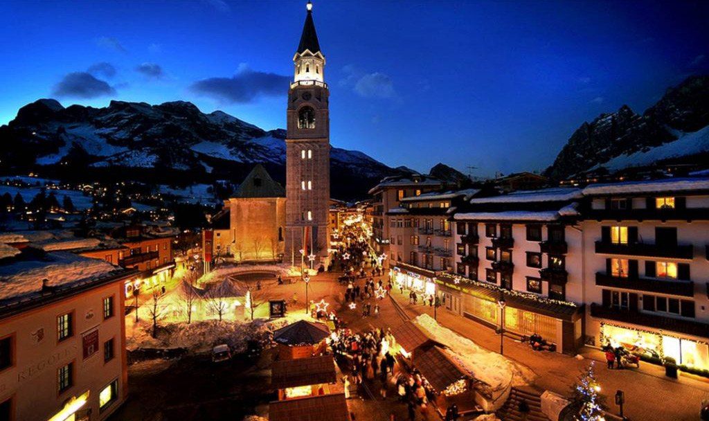 Cortina d’Ampezzo at dusk: the lit bell tower, festive stalls and a busy main street beneath snowy mountains