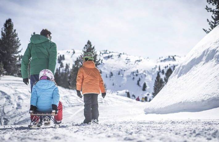 A family on a snowy track: a small child on a wooden sledge, a parent and an older child walking ahead