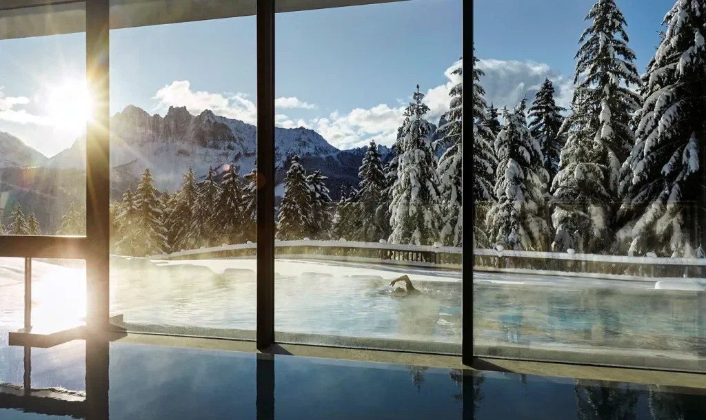 A swimmer in a steaming outdoor pool among snow-laden firs, seen from an indoor pool, with Dolomite peaks in low winter sun