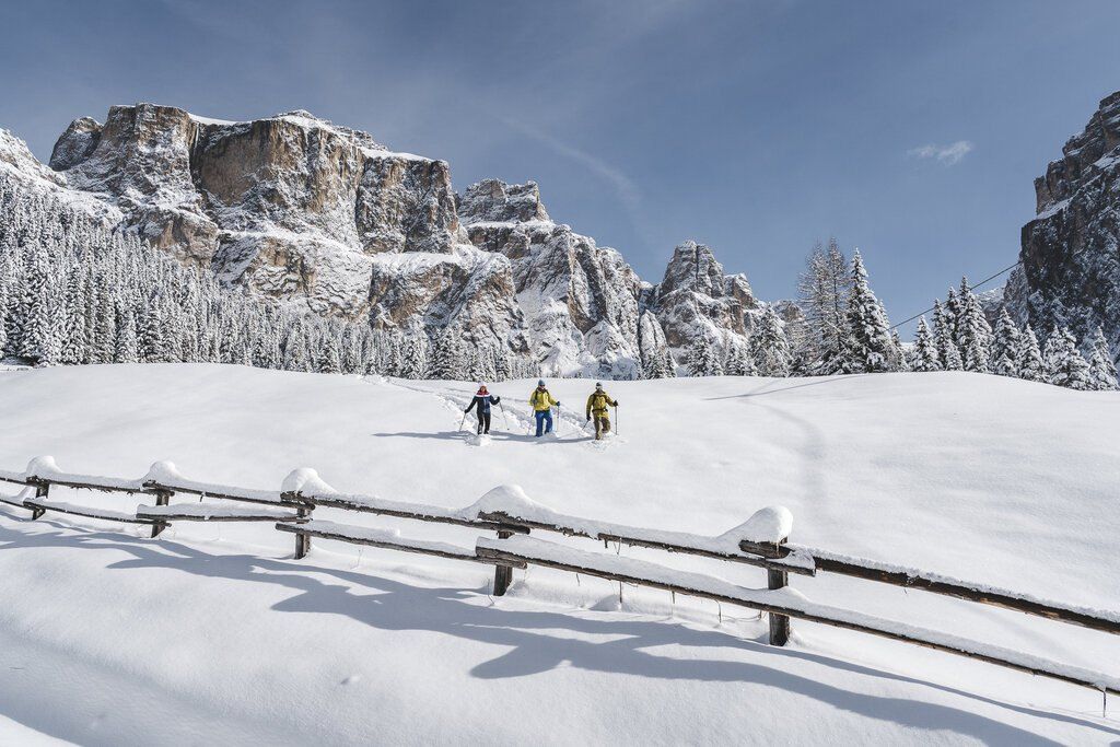Three snowshoers crossing a snowy meadow beyond a wooden fence, below the pale limestone walls of the Dolomites