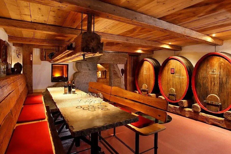 A wine cellar with large wooden barrels, a long stone table laid with glasses and bottles, and red-cushioned benches