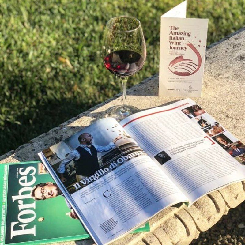 Eine Ausgabe von Forbes und eine aufgeschlagene Zeitschrift auf einer Steinbank, daneben ein Glas Rotwein und das Programm einer Weinveranstaltung